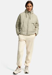 Gevoerde jas in lichtgroen met een hoge kraag, een ritssluiting aan de voorkant en koordjes; gedragen met crèmekleurige sweatpants en lichte sneakers.