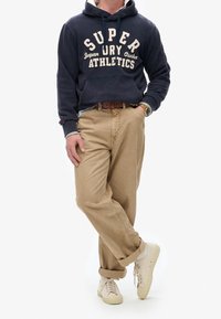 Persoon draagt een marineblauwe hoodie met de tekst 'Superdry Athletics', beige broek, witte sneakers en een polshorloge, staat met gekruiste benen.