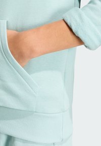 Svetlo mentolová mikina s rebrovaným manžetom, so bočnou kapsou a jemnou textúrou. Materiál sa zdá byť hladký a pohodlný.