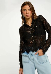 Blusa di pizzo nero con design a bottone, caratterizzata da un orlo ondulato, maniche lunghe e colletto. Abbinata a jeans chiari a vita alta.