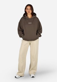 Καφέ oversized φούτερ με μπροστινή τσέπη και λευκό κείμενο "PEGADOR". Συνδυασμένο με ανοιχτό καφέ υφασμάτινο παντελόνι με φαρδιά πόδια και λευκά αθλητικά παπούτσια.