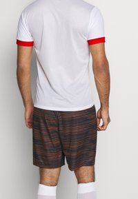 Hvid atletisk trøje i mesh med røde ærmer, kombineret med brune shorts med et horisontalt stribet mønster. Korte hvide sokker er synlige.