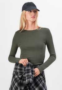 Olijfgroene longsleeve top met een slim fit, geribbelde textuur, gecombineerd met een zwart en grijs geruite hem die om de taille is geknoopt. Pet met logo.