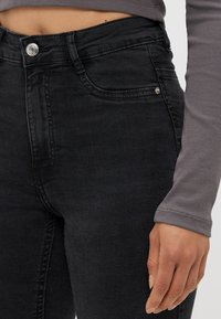 Črne denim skinny jeans z visokim pasom, z zapiranjem na gumb spredaj, podrobnostmi žepov in gladkim, nekoliko teksturiranim materialom.
