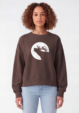 Junge Frau mit lockigem Haar trägt einen braunen Pullover mit einem weißen, runden Design eines Hirsches mit Geweih.