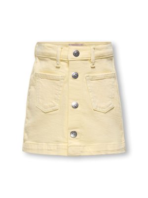 Jeune fille debout portant un t-shirt violet avec un motif de cornet de glace fleuri, une jupe jaune pâle avec des boutons sur le devant et des baskets blanches.