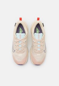 Baskets de sport beige clair avec un upper en maille, accents violets et semelle texturée. Présentent une languette bleue et une boucle de talon rouge pour un enfilage facile.