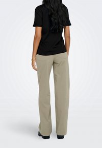 Femme aux longs cheveux foncés portant un t-shirt noir, un pantalon beige à jambes larges, des chaussures noires et un bracelet en or, debout de dos.