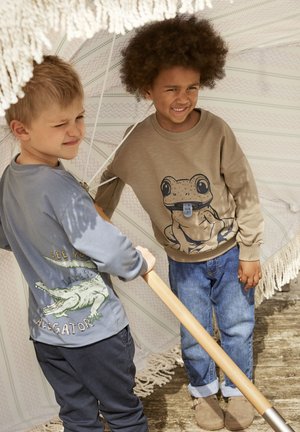 Due bambini si trovano sotto un grande ombrello. Uno indossa una maglietta a maniche lunghe blu chiaro con una grafica di coccodrillo, mentre l'altro indossa un felpa beige con un design di rana. Entrambi indossano pantaloni in denim.