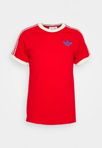 Červené tričko s krátkým rukávem, s krémovým žebrovaným límcem a manžetami. Obsahuje modrou vyšívanou nášivku na hrudi a bíl é pruhy na ramenou.