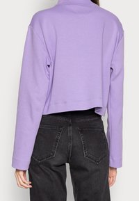 Sudadera corta de color lavanda con mangas largas, hecha de una tela suave, con un cuello redondo y un dobladillo limpio por encima de unos jeans grises de talle alto.