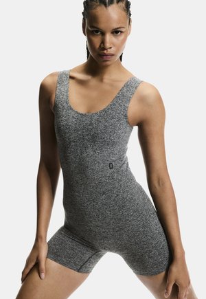 Ženska v prilagojenem sivem športnem igralnem kombinezonu brez rokavov, s pletenimi lasmi, stoji razprtih nog in z rokami na stegenih na belem ozadju.