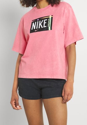Rosa, übergroßes Baumwoll-T-Shirt mit einem schwarzen Nike-Logo-Graphic auf der Vorderseite. Kurze Ärmel und eine lockere Passform. Kombiniert mit dunklen Shorts.