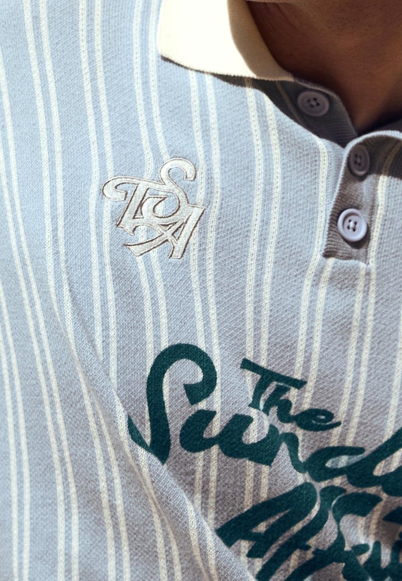 Gros plan d’un polo rayé bleu clair et blanc avec un col blanc, un logo brodé et un texte imprimé en vert.