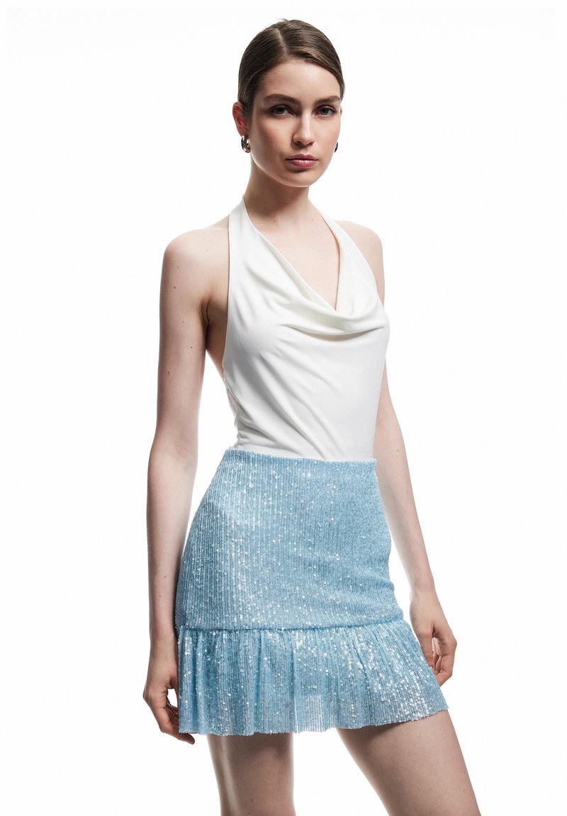 Jeune femme aux cheveux bruns portant un top dos nu blanc et une jupe bleu clair à sequins avec un ourlet volanté, debout devant un fond blanc.
