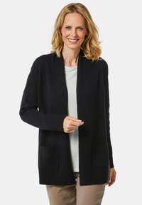 Schwarzer Cardigan mit offenem Vorderteil, gerippten Bündchen und zwei Fronttaschen, aus einem weichen Material, über einem hellen Hemd getragen.