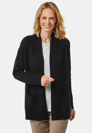 Cardigan noir à devant ouvert, avec des poignets côtelés et deux poches avant, fabriqué dans un matériau doux, porté sur une chemise légère.