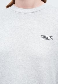 Primer plano de una camisa gris claro con "ROSIE" y el logo de Puma impresos en negro en la parte superior del pecho cerca del collar.