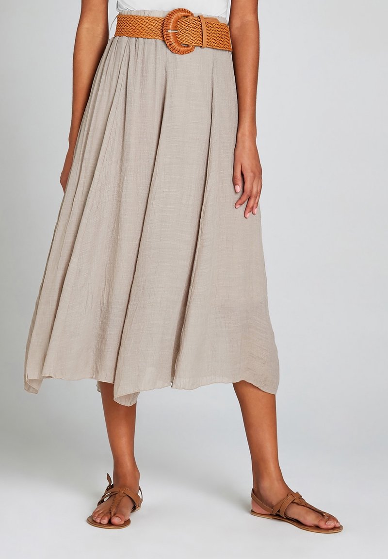 Femme portant une jupe midi beige avec une large ceinture tissée marron et des sandales plates marron, debout devant un fond clair uni.