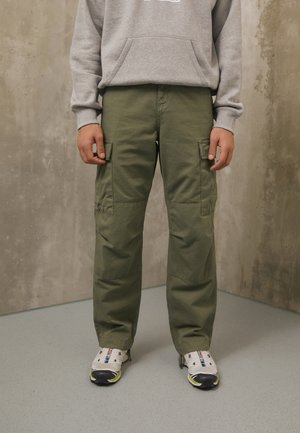 Person wearing olive green cargo pants, gray sweatshirt, and white athletic shoes standing on a gray floor against a textured wall.