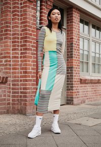 Robe à manches longues présentant un design patchwork en jaune, bleu sarcelle et crème avec des rayures horizontales, complétée par des baskets blanches et un sac bleu sarcelle.