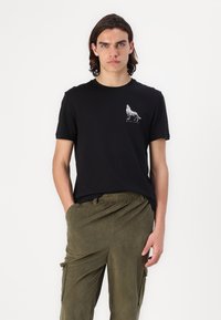Zwarte katoenen t-shirt met een witte wolfafbeelding op de linkerkant van de borst. Gecombineerd met olijfgroene ribgebreide cargobroek.