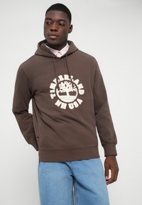Jauns vīrietis, kurš valkā brūnu Timberland kapuci virs rozā krekla ar apkakli un gaiši ziliem džinsiem, skatās uz sāniem uz vienkārša fona.