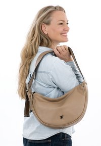 Blond naeratav naine, kes kannab heledat teksasärki ja kandes suurt beežikat poolkuu kujuga õlakotti.