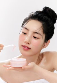 Pot de patchs pour les yeux au rétinol Abib, en plastique rose avec couvercle transparent, tenu par une personne appliquant un patch en gel sous l'œil.