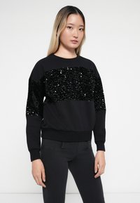 Sweatshirt preto com um painel de lantejoulas ao longo do peito e das mangas. Apresenta decote redondo e punhos e bainha canelados. Textura suave em geral.