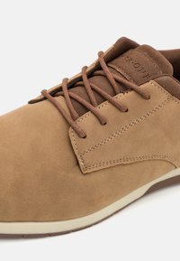 Chaussure en suède marron avec un bout arrondi, un laçage à l'avant et des lacets marron contrastés. Présente une surface lisse et texturée ainsi qu'une semelle légère.