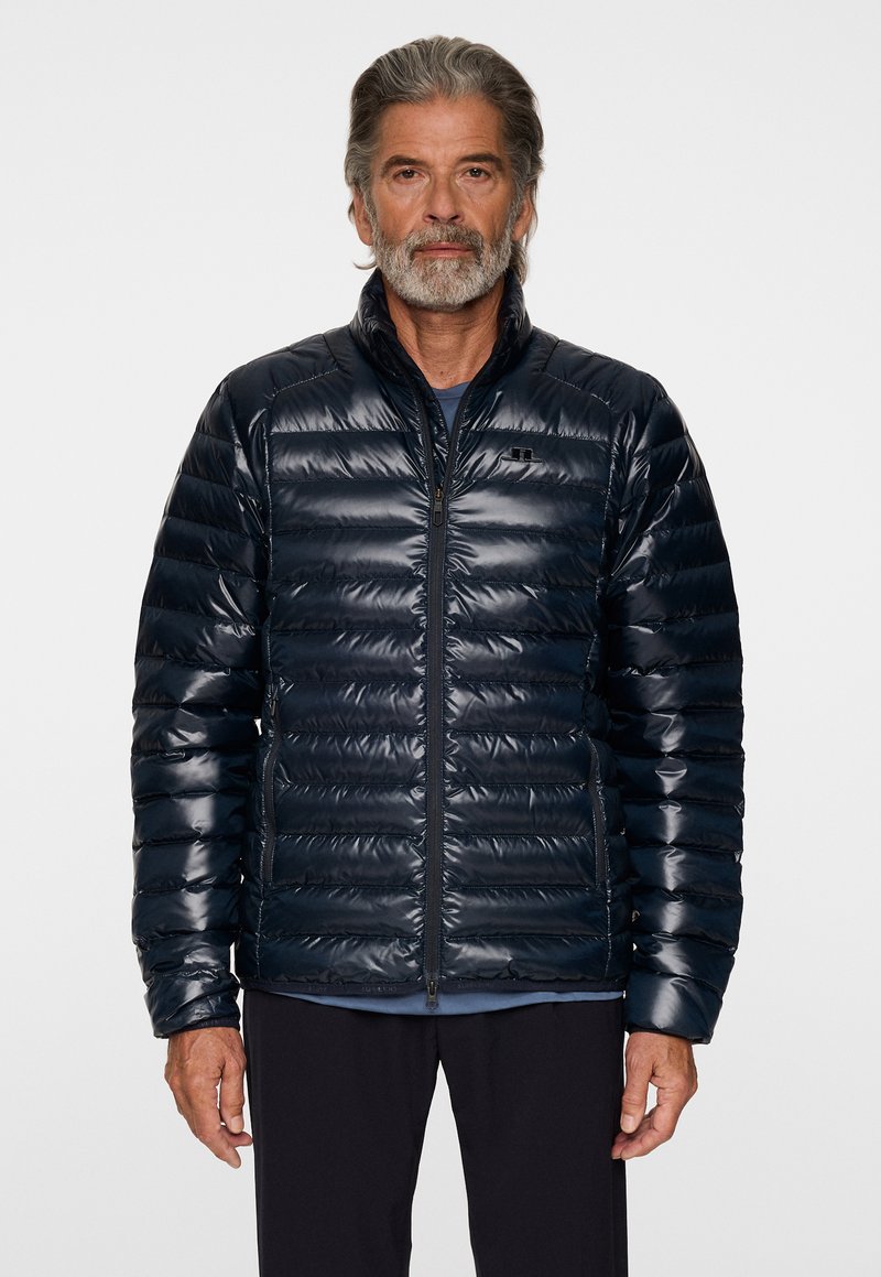 Tummansininen puhvitakki kiiltävällä pinnalla, tikattu muotoilu, korkea kaulus, etuvetoketju ja raglan-hihat. Sopii ulkoiluun.