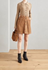 Top de punto acanalado beige con botones en la parte delantera, combinado con una falda de pana marrón con bolsillos y botones, complementado por botas negras hasta el tobillo.