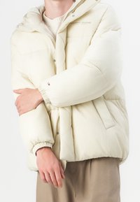 Prošívaná krémová bunda s kapucí, elastickými manžetami a zapínáním na patentky. Na hrudi a po stranách kapsy je logo. Texturovaná látka.