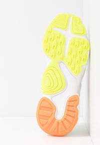 Sula av Adidas-sneaker med gula, orangea och vita gummimönster, placerad vertikalt mot en vit bakgrund.