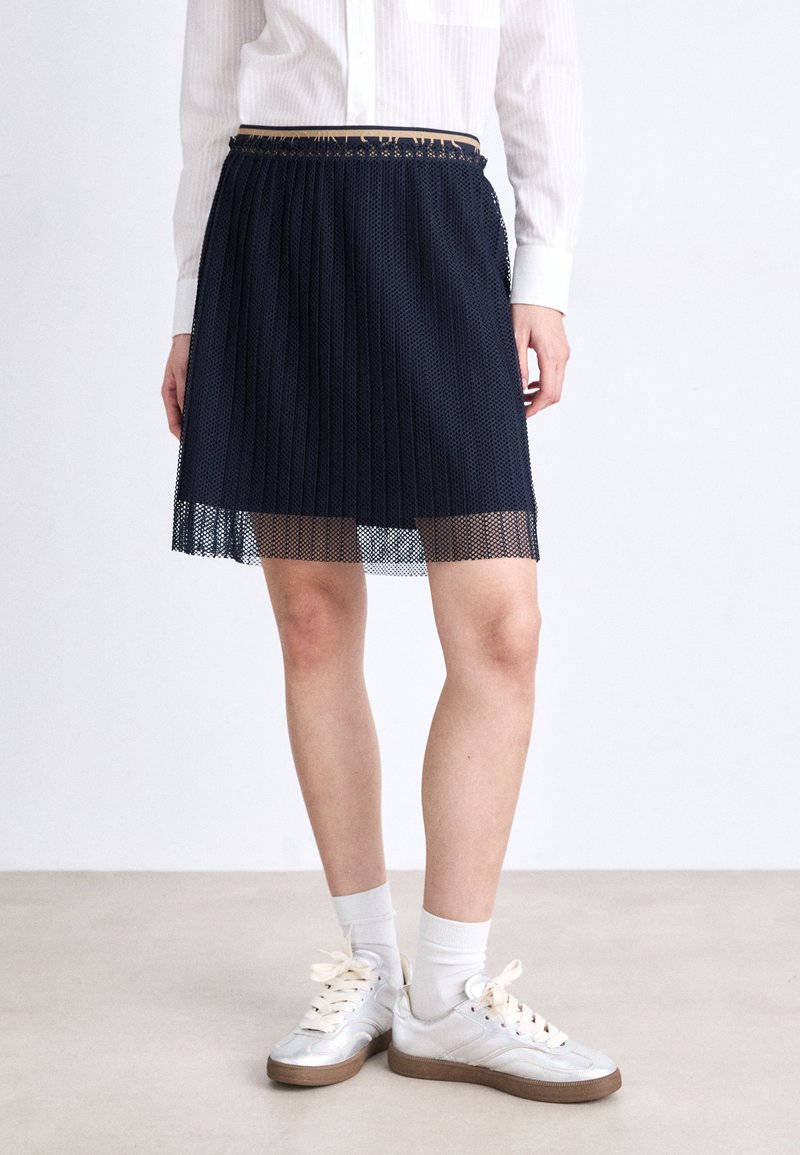 Falda de marine con plisados y un dobladillo de malla transparente, combinada con zapatillas plateadas y calcetines blancos, sobre un fondo neutro.
