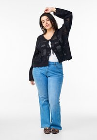 Svart stickad cardigan med öppen väv, vågiga kanter och knytning framtill, matchad med ljusblå utsvängda jeans och flats i leopardmönster.