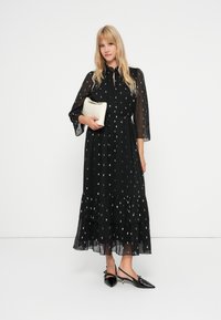 Mustat pitkät hihansuut omaava mekko, jossa on kerroksellinen röyhelöhelma, hopeapistekuvio, nappisulku ja läpikuultava kangas. Yhdistetty kiiltävien mustien avokkaiden ja clutch-laukun kanssa.