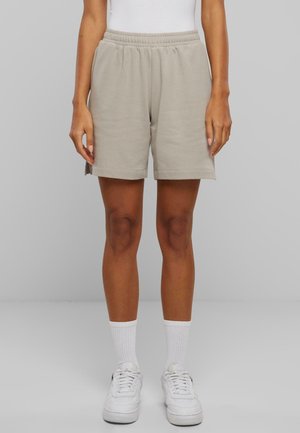 Personne portant un short beige à taille élastique, des chaussettes blanches et des baskets blanches, debout devant un fond gris clair uni.