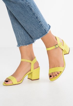 Gelbe Wildleder-Sandaletten mit Blockabsatz, breiten Riemen, Knöchelschnalle und offenen Zehen. Kombiniert mit Jeans mit ausgefranstem Saum, was einen lässigen Stil zeigt.