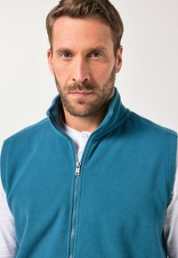 Fleece-Weste in Teal mit hohem Kragen und durchgehendem Reißverschluss. Getragen über einem weißen Langarmshirt. Glatte Textur, keine sichtbaren Muster.