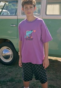 Paarse katoenen t-shirt met een kameleonafbeelding en de tekst "KEEP ROLLIN'", gecombineerd met zwart en grijs geblokte shorts. Groene vintage bus op de achtergrond.