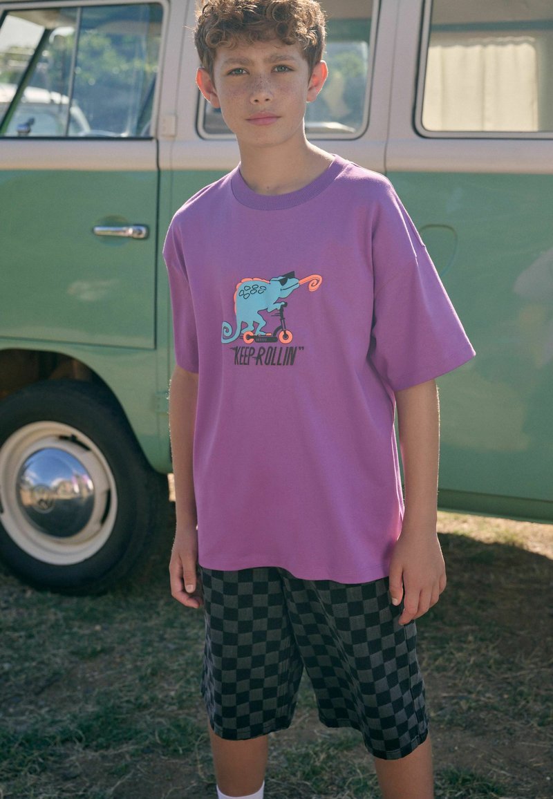 Lila Baumwoll-T-Shirt mit Chamäleon-Grafik und dem Text "KEEP ROLLIN'", kombiniert mit schwarz-grauen karierten Shorts. Im Hintergrund ein grüner Vintage-Bus.