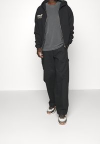 Black zip-up hoodie with side pockets, grey t-shirt underneath, black wide-leg pants, and multicolored sneakers with white soles.