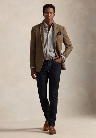 Mand iført brun blazer, lysegrå skjorte, mørke jeans, brun bælte og sko, briller, med den ene hånd i lommen mod ensfarvet baggrund.