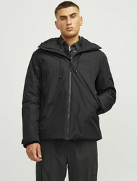 Chaqueta negra impermeable con capucha, cremallera frontal, cordones ajustables y un forro interior texturizado, llevada sobre una camisa de estampado.