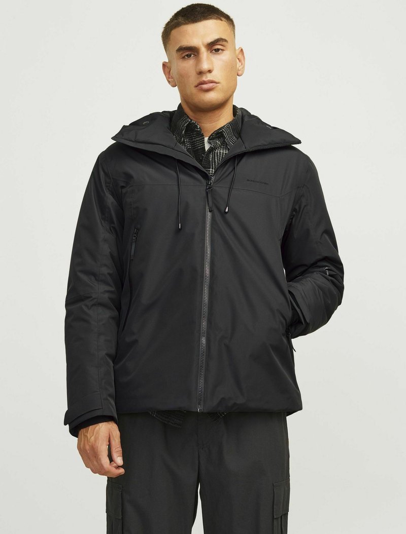 Chaqueta negra impermeable con capucha, cremallera frontal, cordones ajustables y un forro interior texturizado, llevada sobre una camisa de estampado.