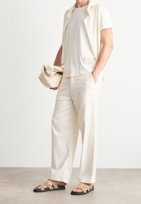 Kostym i krämfärg med en t-shirt med korta ärmar, vida byxor och sandaler med remmar. Accessoarpåse i handen, minimalistisk design.