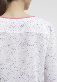 Weiße Bluse mit hellblauem Punktmuster, runder Ausschnitt mit rosa Saum, plissierte Ärmel und Strukturstoffdetails. Rückansicht.