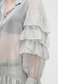 Διαφανές γκρι πουκάμισο με στρωματοποιημένα μανίκια με βολάν και μανσέτες με κουμπιά, φορεμένο πάνω από μαύρο εσώρουχο, εμφανιζόμενο από τους ώμους έως τη μέση.